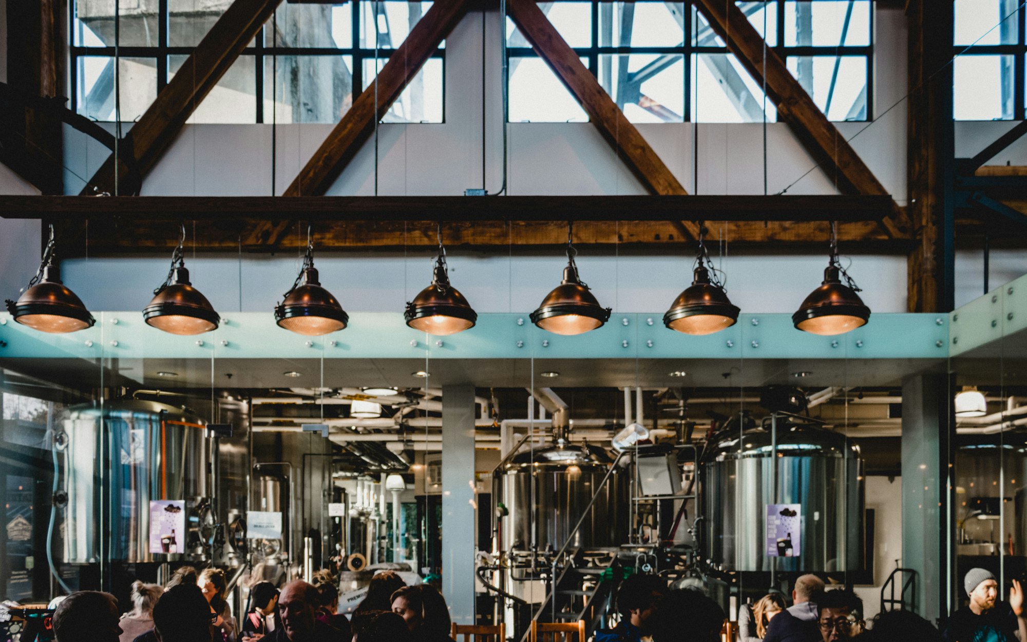 The taproom under the brewery roof trusses, tanks behind the glass wall and lamps over the tables